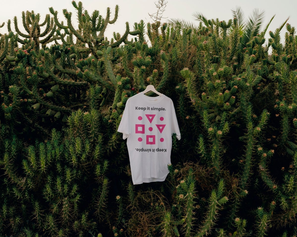 Wit T-shirt met de logovormen in magenta en de tekst Keep it simple, hangend voor een cactushaag